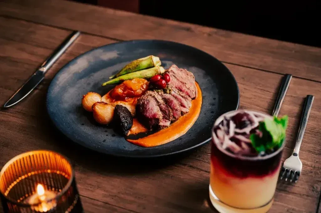 Gourmet meat dish, vegetables, layered cocktail, and candle on dark wood.