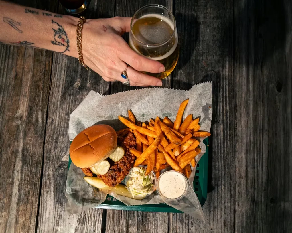 Fried chicken sandwich, fries, coleslaw, and frothy beer on rustic table.