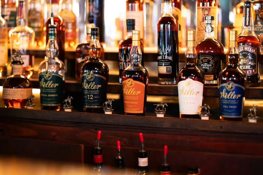Well-stocked bar display of Weller bourbon bottles on wooden shelves.