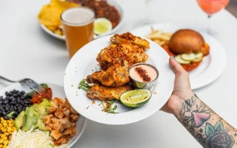 Tattooed hand holding spiced chicken wings with a spread of food and drinks.