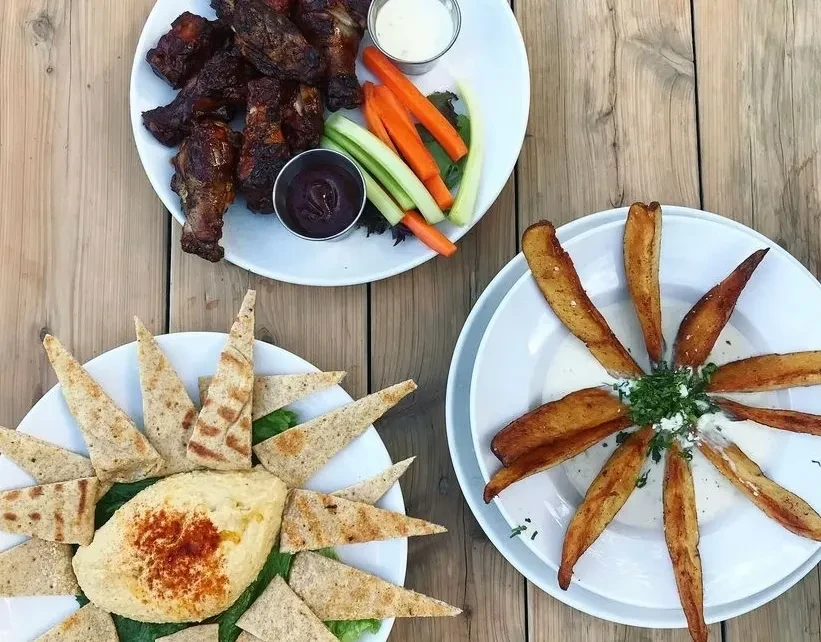 Overhead of chicken wings, hummus, and potato wedges on a wooden table.