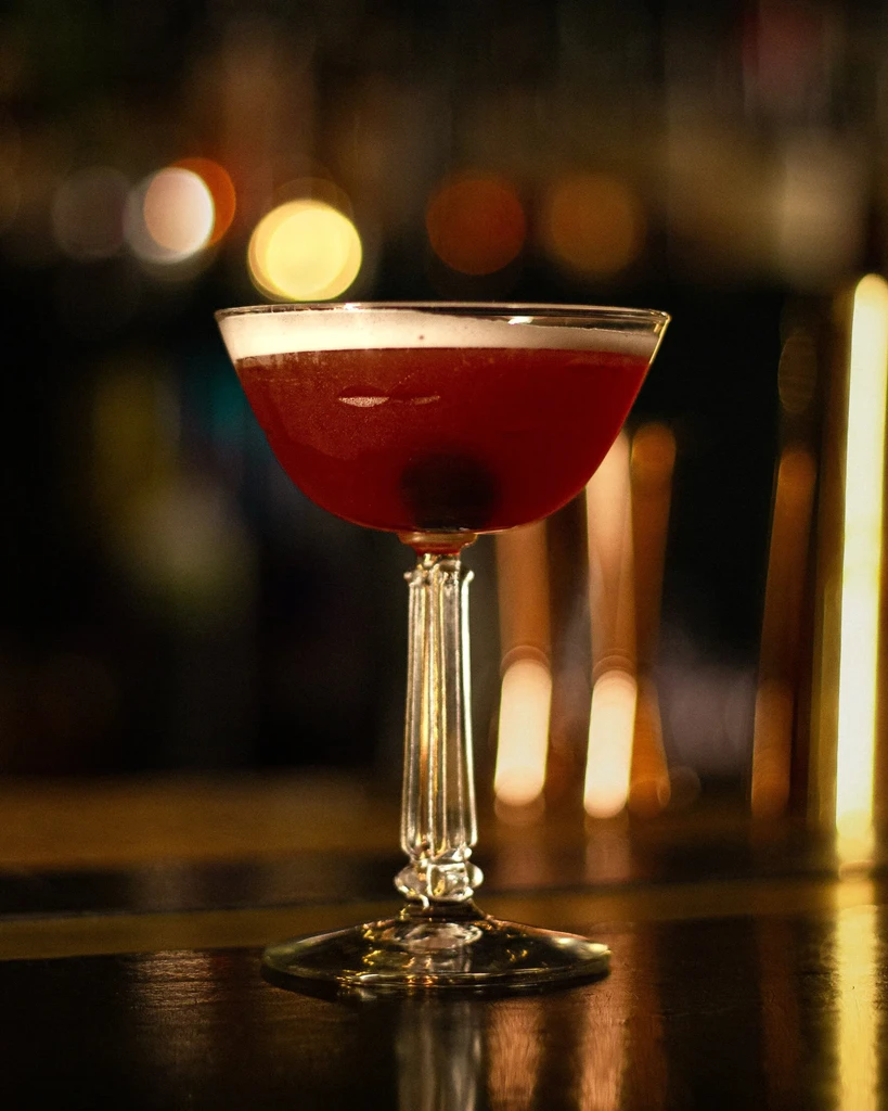 Dark red cocktail with white foam and cherry in ornate coupe glass.