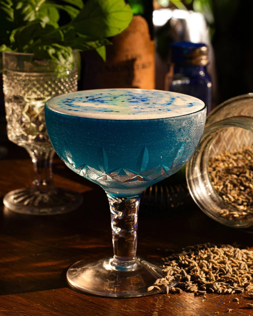 Vibrant blue cocktail in cut glass, with frothy top and lavender.