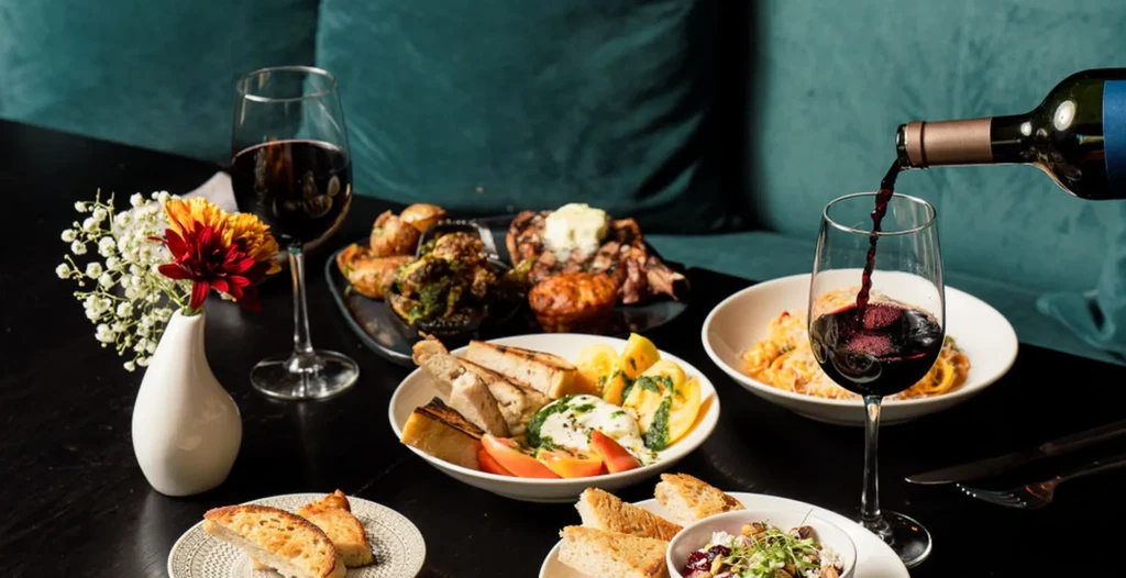 Dining table with diverse dishes, red wine pouring, and decorative flowers.
