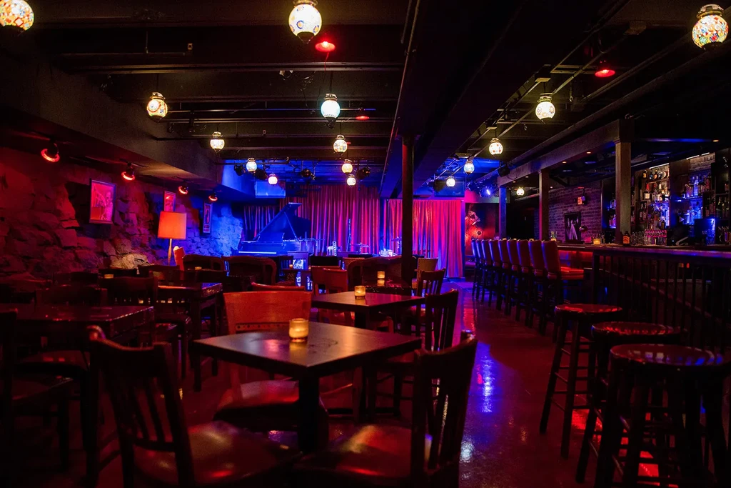 Empty, atmospheric bar with stage, grand piano, and dramatic red-blue lighting.