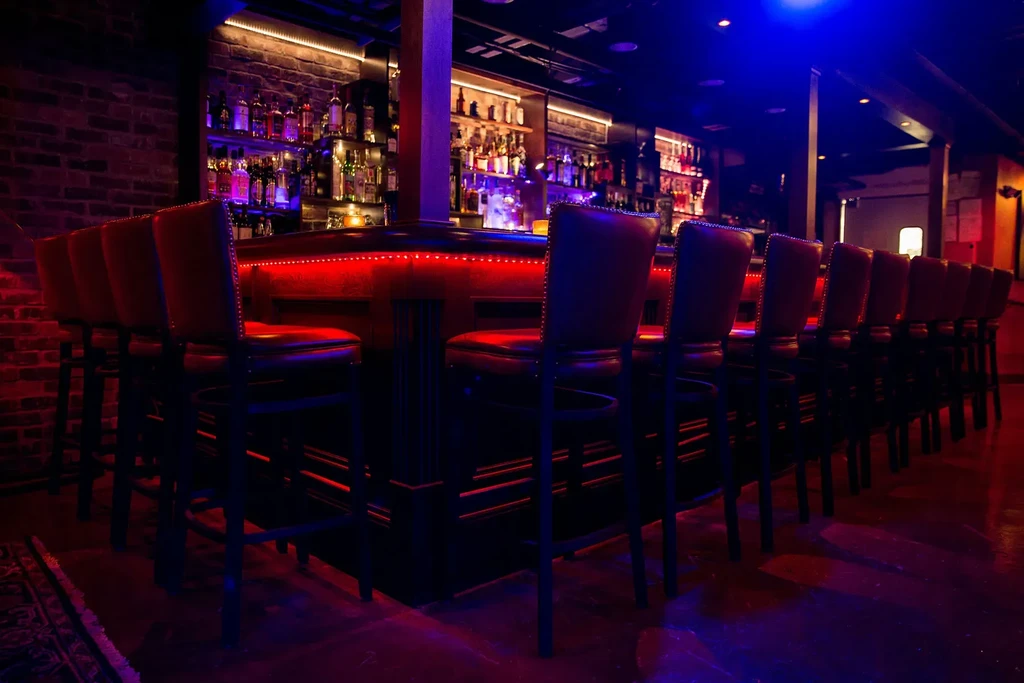 Dimly lit bar counter with red LED, empty stools, liquor bottles.