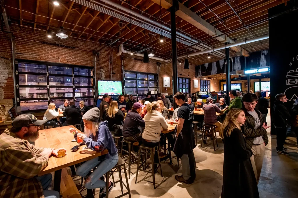 Bustling industrial-chic bar with patrons socializing and watching sports.