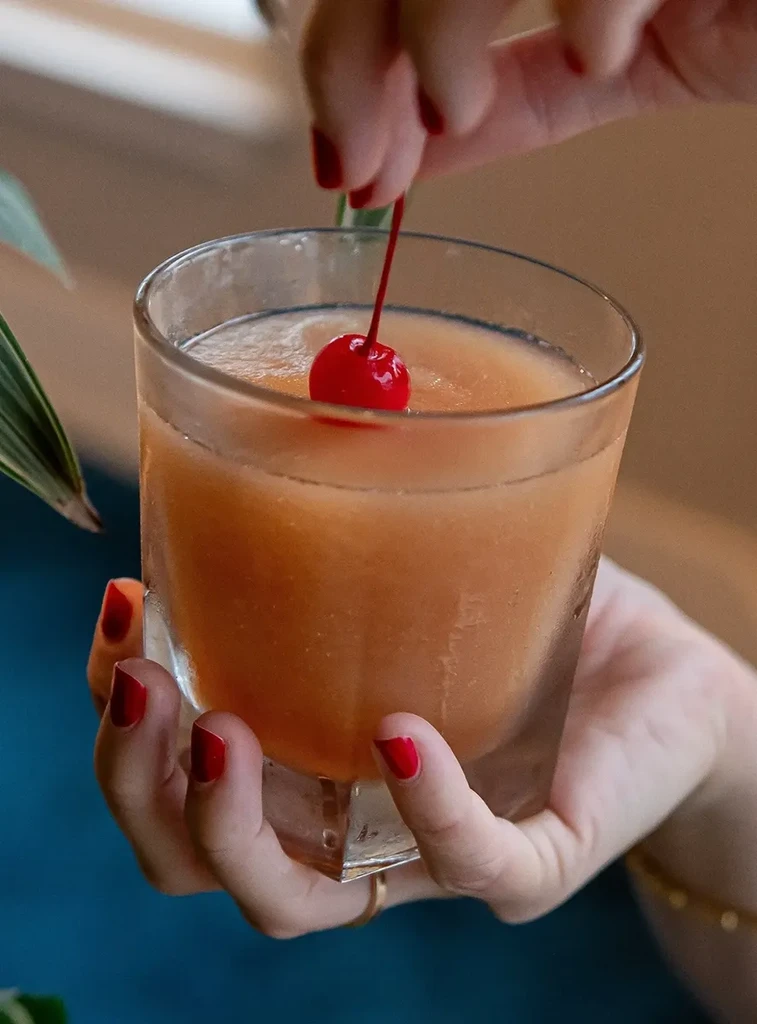Red-nailed fingers add a maraschino cherry to an orange blended drink.