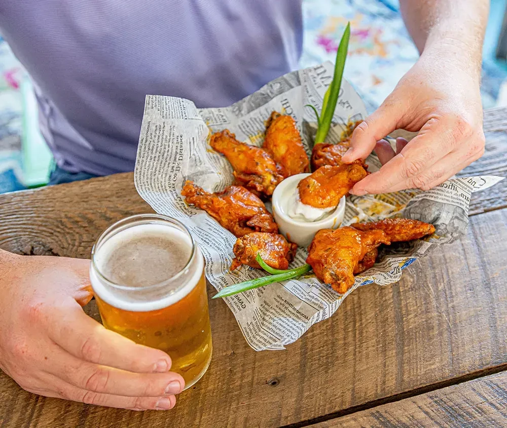 Hands dipping buffalo wings into sauce, holding beer on wood table.