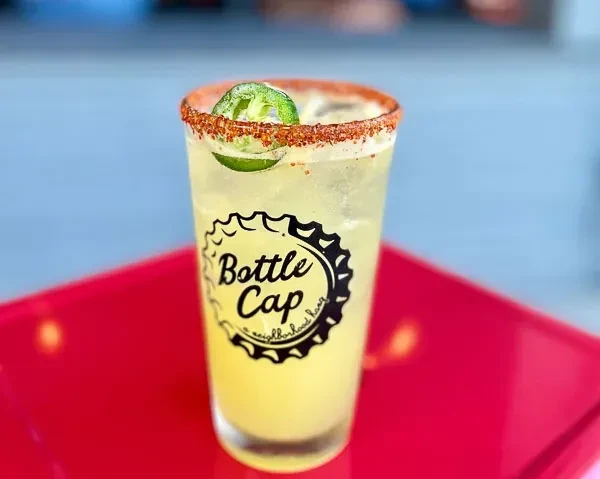 Pale yellow-green cocktail with a spiced rim, jalapeño slice, and bottle cap logo.