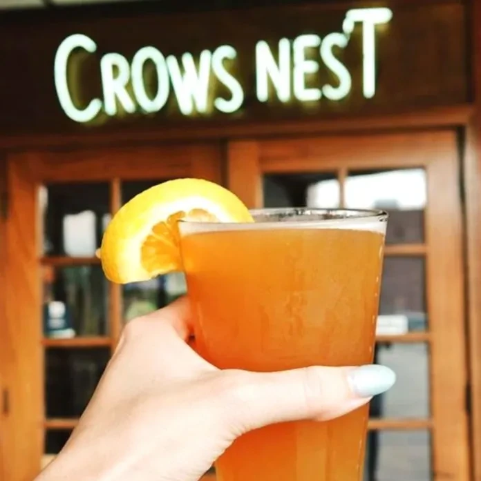 Hand with blue nails holds amber drink, lemon garnish, 'CROWS NEST' sign.