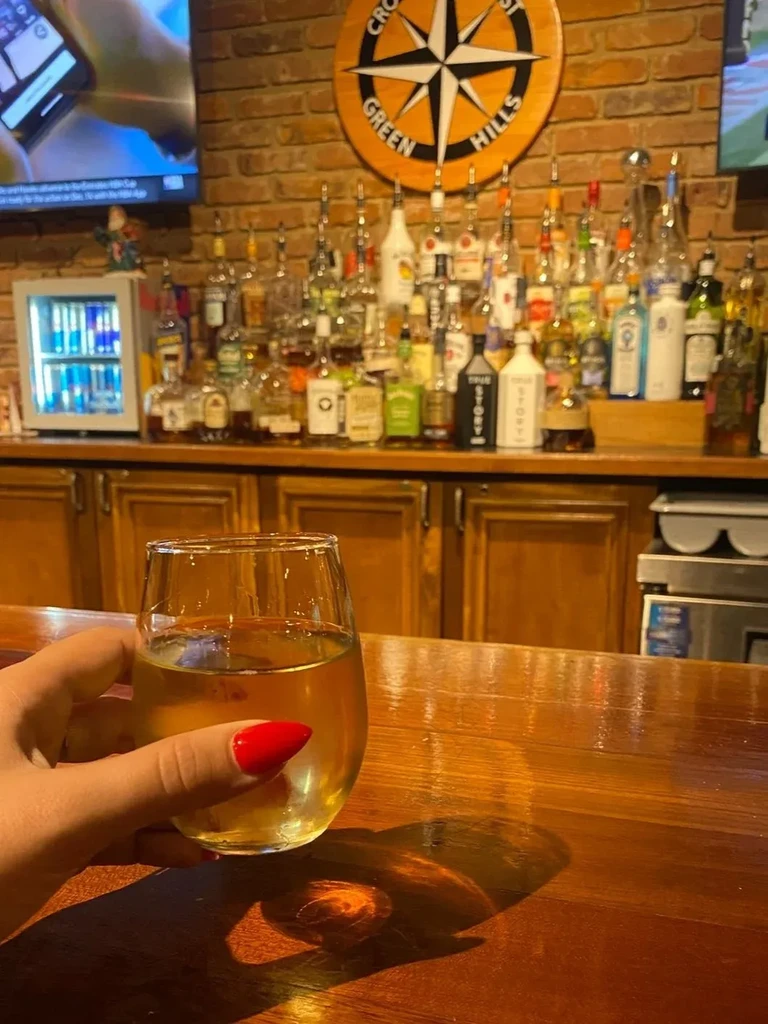 Hand with red nails holds drink on bar, "CROSS GREEN HILLS" sign.