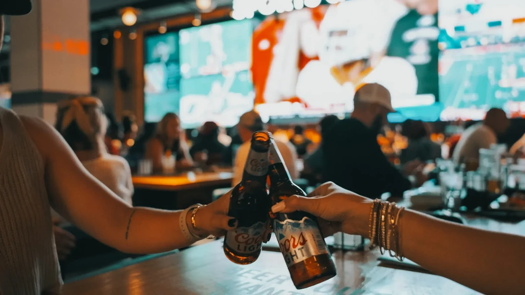 Two hands clinking Coors Light bottles in a blurred sports bar.
