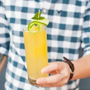 Person in checkered shirt holds yellow cocktail with cucumber and mint.