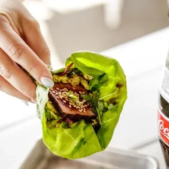 Hand holds green lettuce wrap with seared meat, sesame seeds, herbs.
