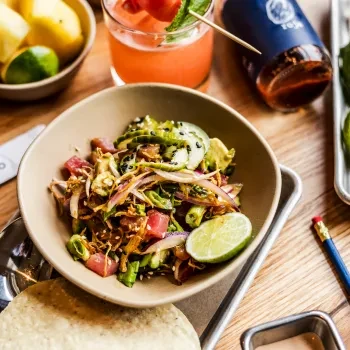 A vibrant poke bowl, cocktail, and limes arranged on a wooden table.