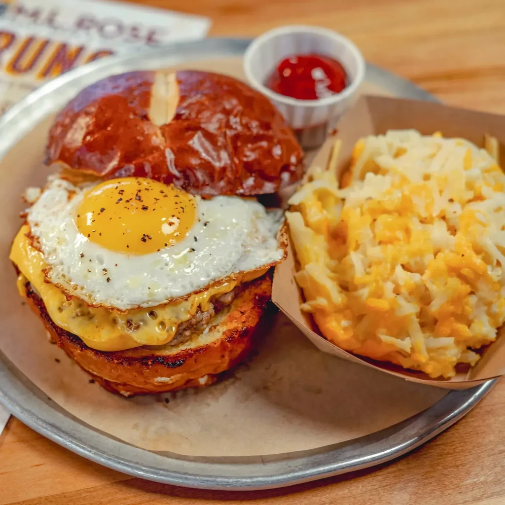 Breakfast burger with fried egg, hash browns, ketchup, on a metal tray.