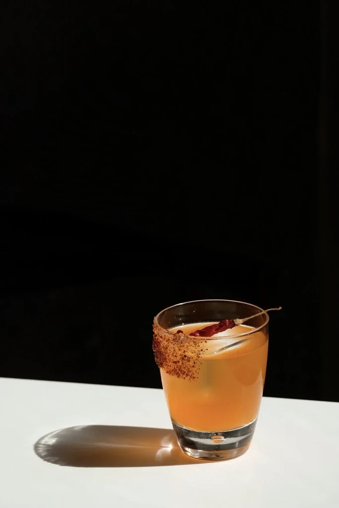 Amber cocktail in tumbler glass, spice rim, dried chili garnish.