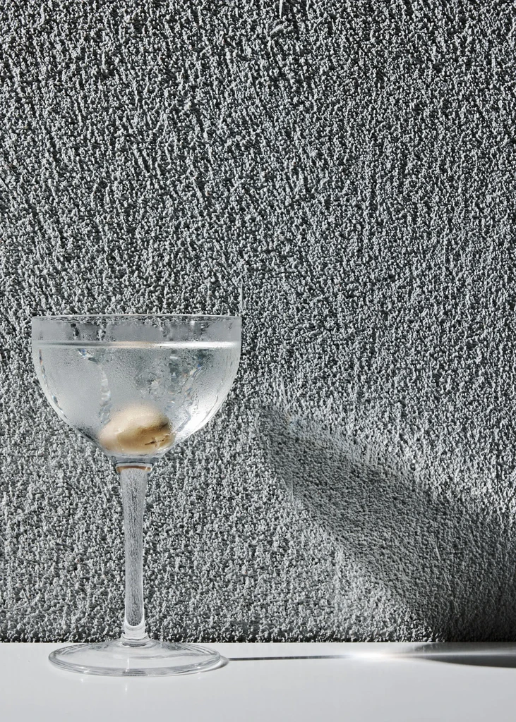 Chilled cocktail glass with clear liquid, green olive, dark textured background.