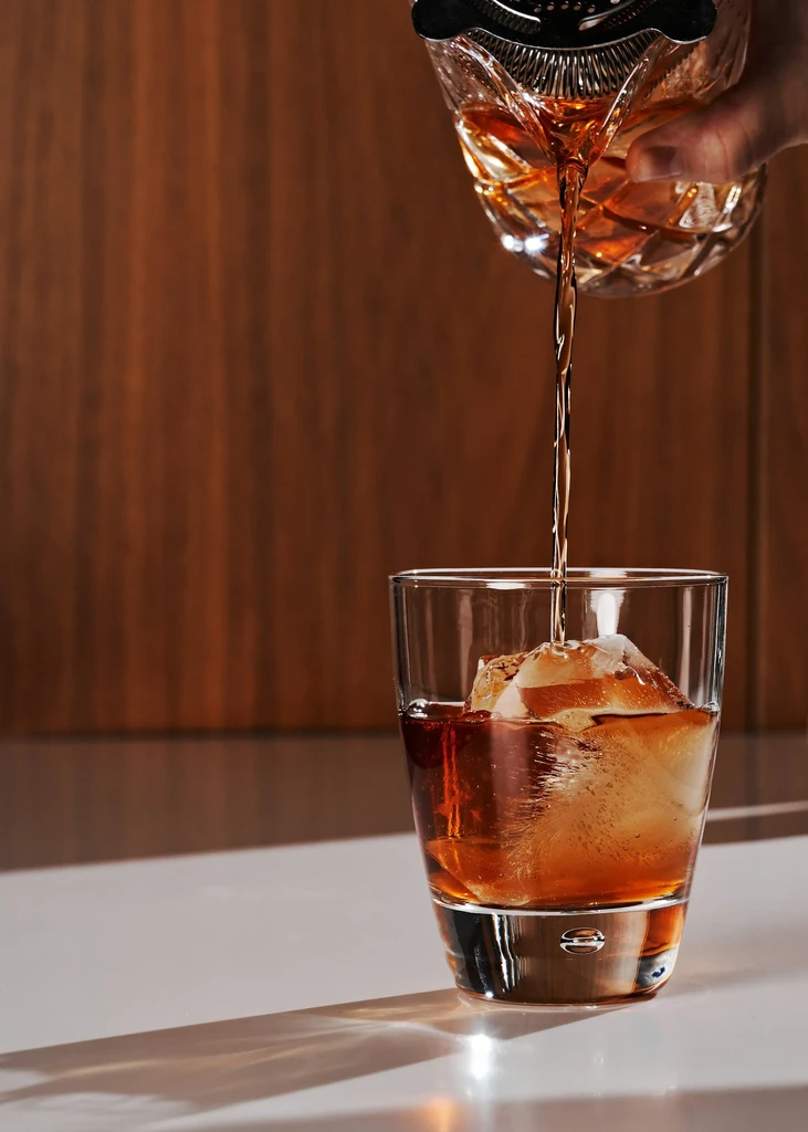Dark amber liquid pours through strainer into rocks glass with ice.