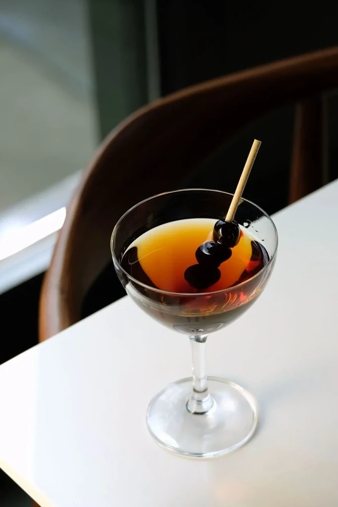 Dark amber cocktail with two maraschino cherries in a coupe glass.