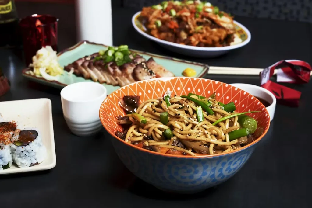Variety of Asian dishes: colorful noodles, sushi rolls, and sliced pork.