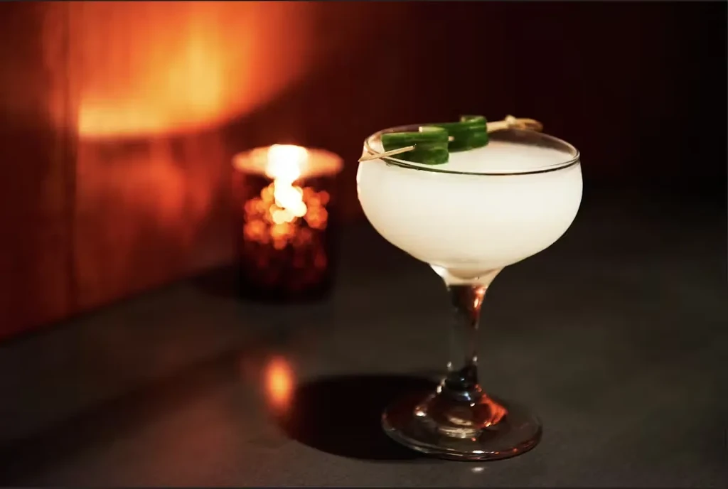 Creamy white cocktail in coupe glass with green garnish and warm candle.