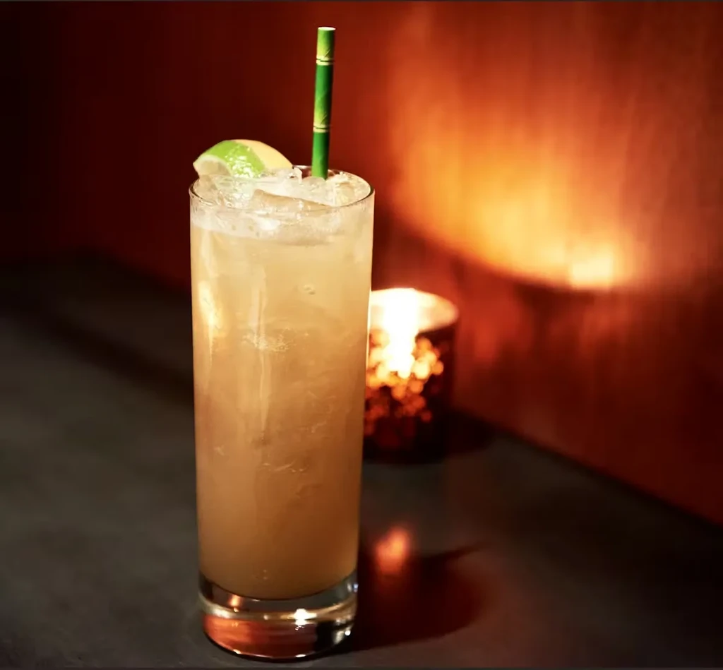 Amber cocktail with lime and straw, candlelit on a dark surface.