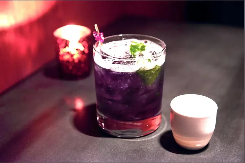 Purple cocktail, frothy top, mint, pink pick, candle, and cup.