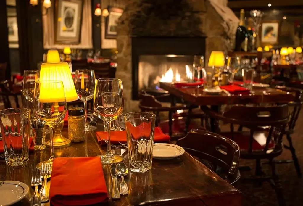 Dimly lit restaurant tables set for dinner with a burning fireplace.