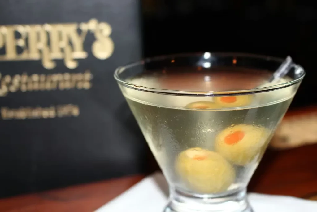 Chilled amber martini with three pimento olives and blurred restaurant sign.