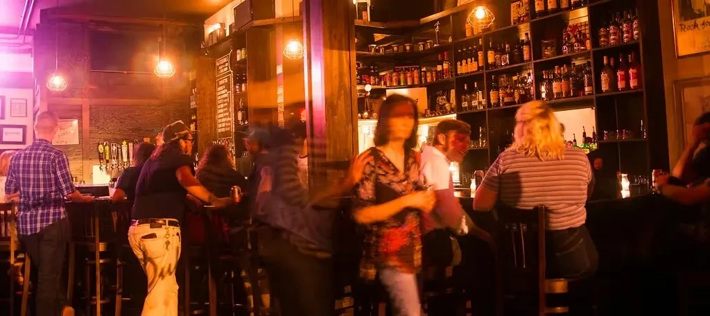 Warmly lit bar interior with a long wooden counter and indistinct patrons.