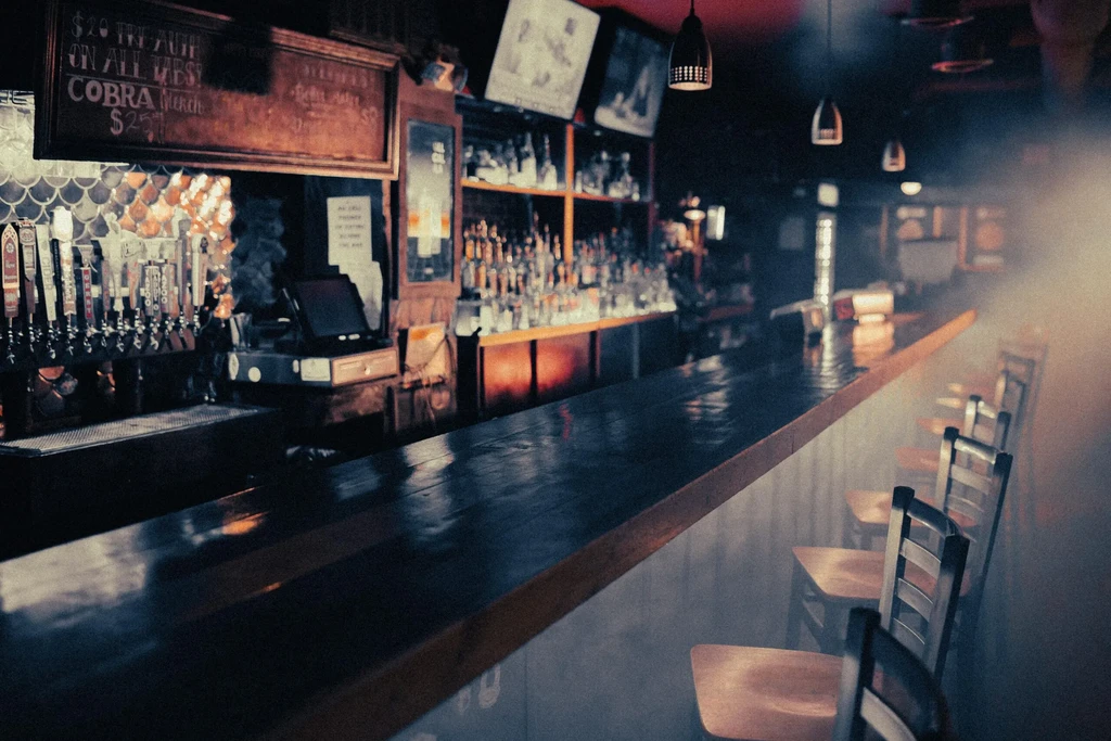 Dark wooden bar, empty stools, beer taps, liquor bottles, and rustic sign.
