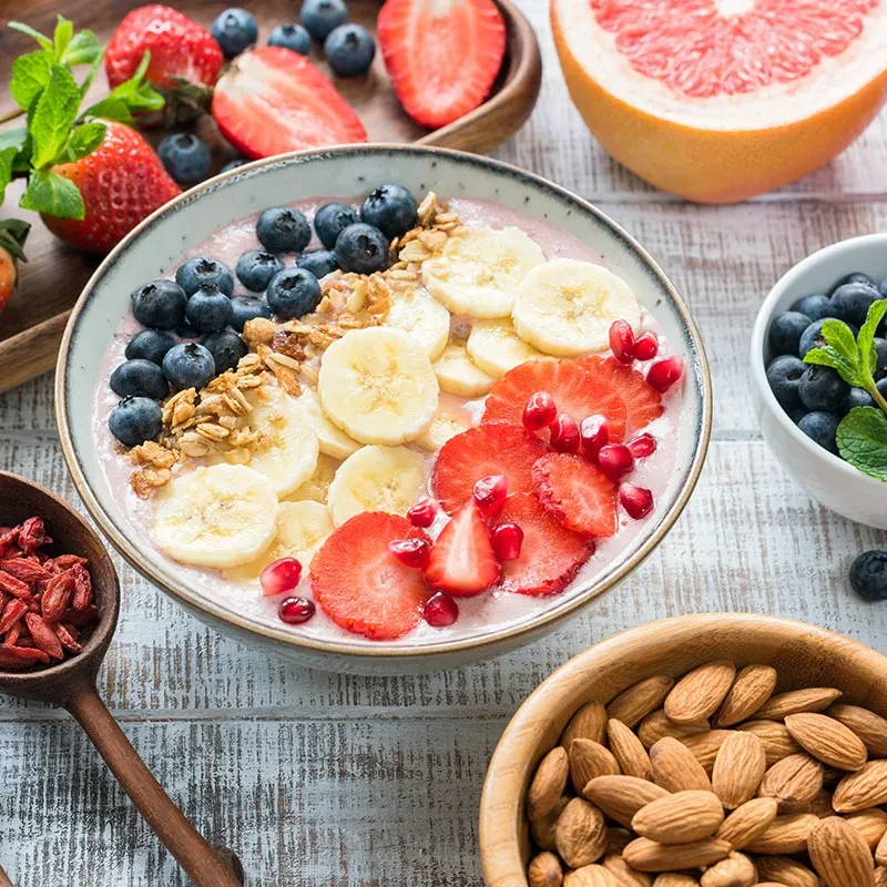 Smoothie bowl topped with berries, banana, granola, and almonds on wood.