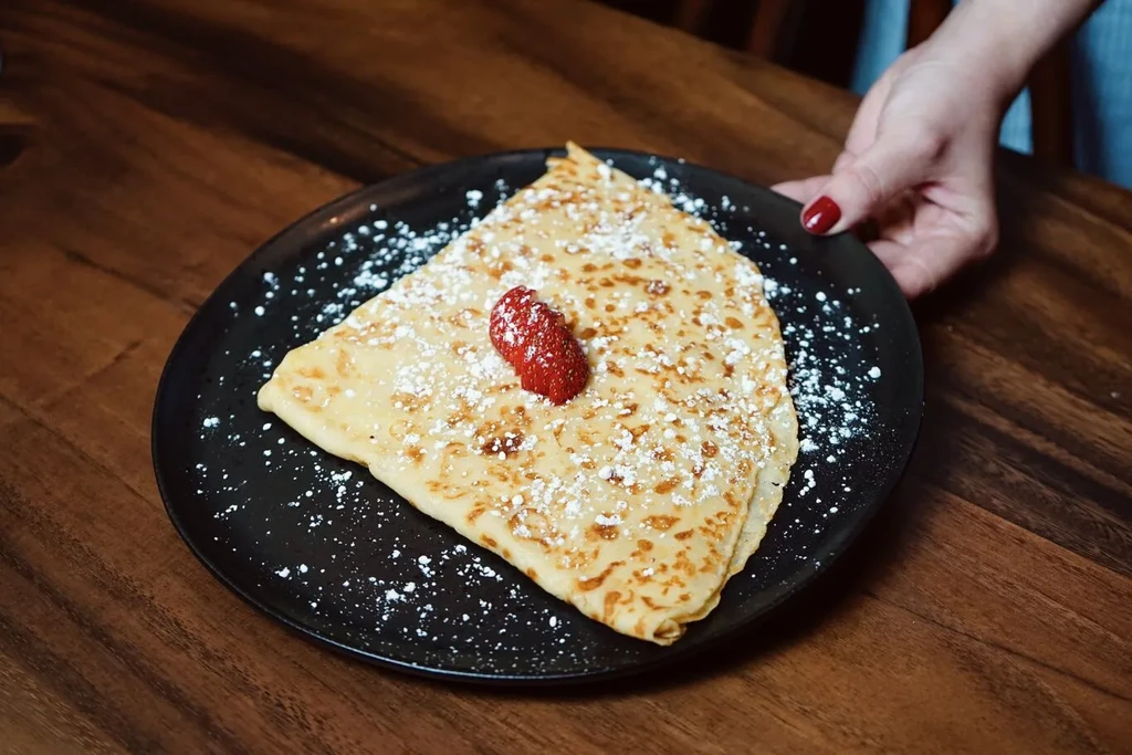 Hand holding a triangular, sugar-dusted crepe with strawberry on a black plate.