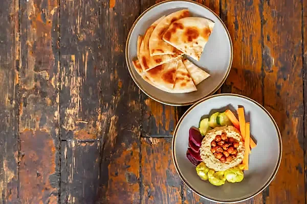 Hummus with chickpeas and vegetables beside charred pita on wood table