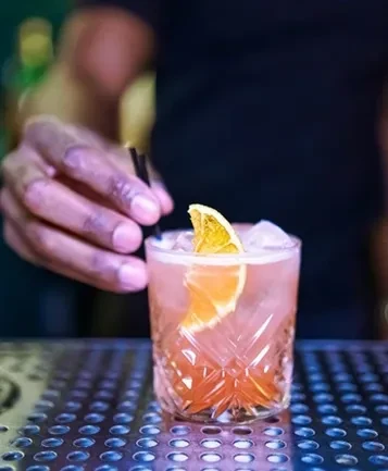 Pink-orange cocktail with ice and dried orange slice, hand adding straw.