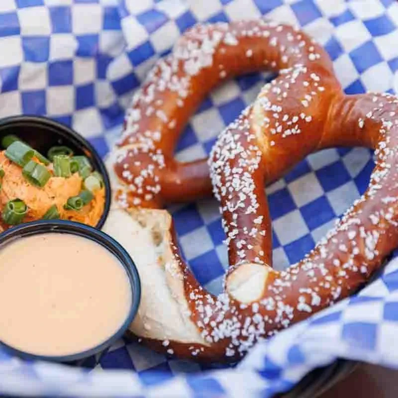 Two salted soft pretzels in a basket with checkered paper and two dips.
