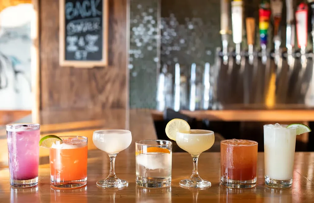 Seven distinct cocktails with varied garnishes on a wooden bar.