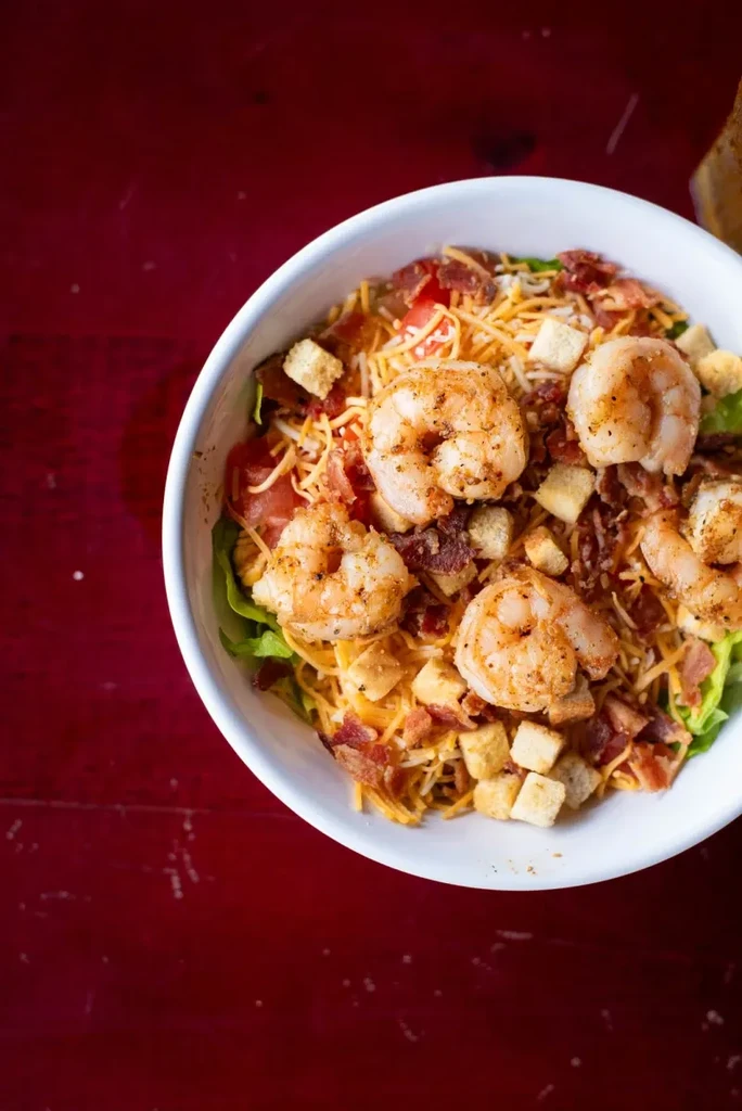 Vibrant shrimp salad with cheese, bacon, croutons, tomatoes in white bowl.