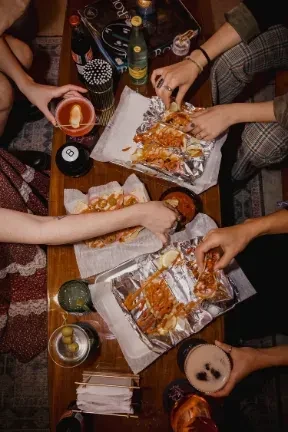 Overhead shot: hands sharing grilled food and cocktails on a wooden table.