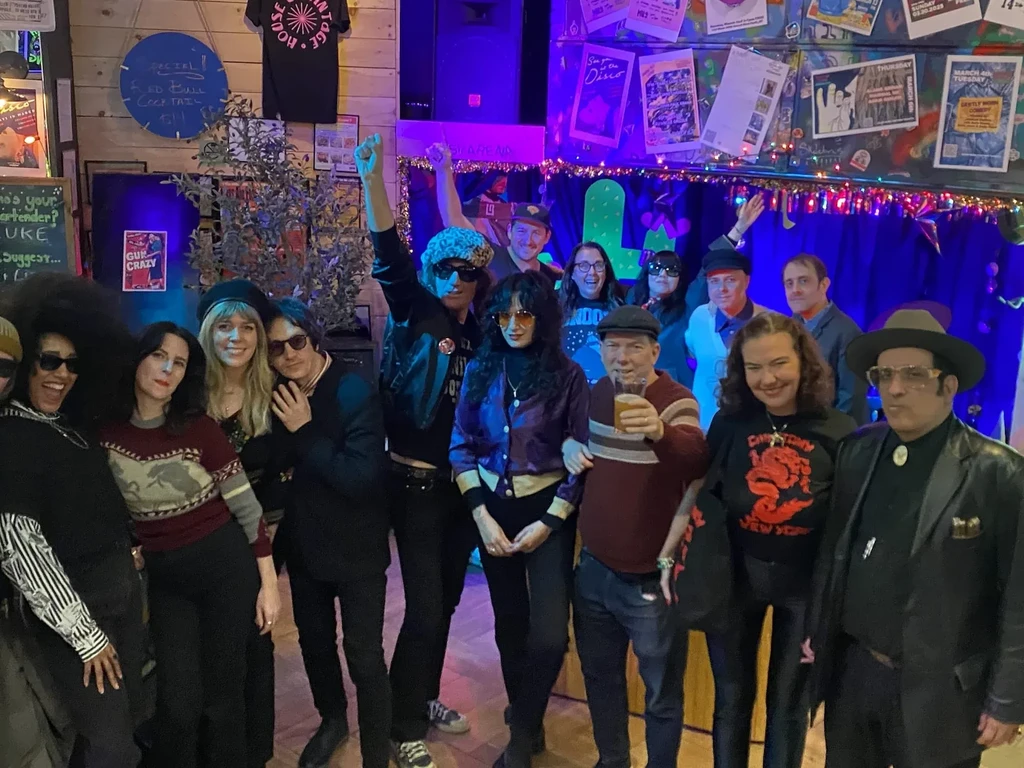 Smiling group of 15 people posing in a dark, poster-filled bar.