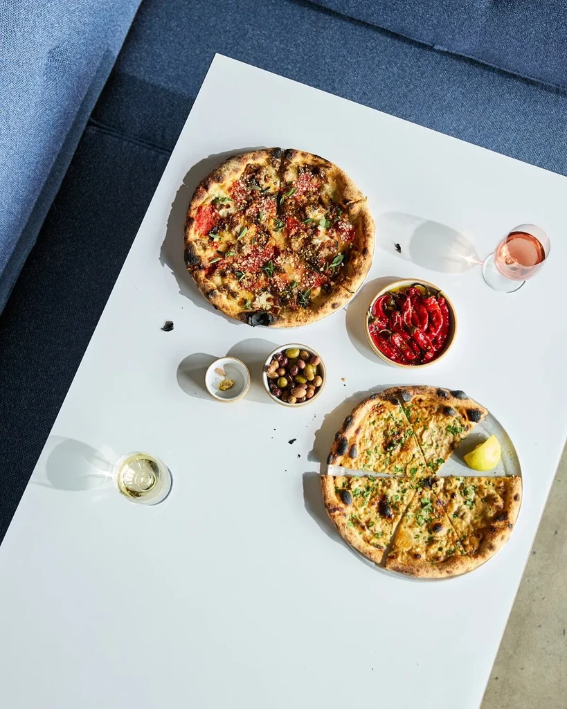 Pizzas, olives, roasted peppers, and wines on a bright white table.