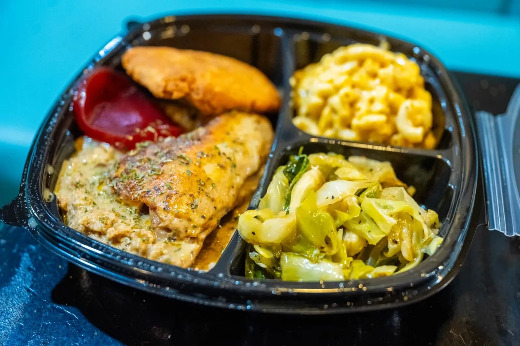 Black take-out container with chicken, mac and cheese, cabbage, and a fritter.