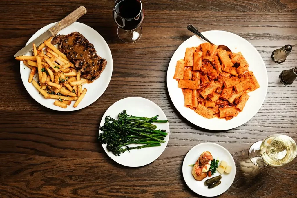 Steak, pasta, vegetables, appetizer, and wine laid on a dark table.