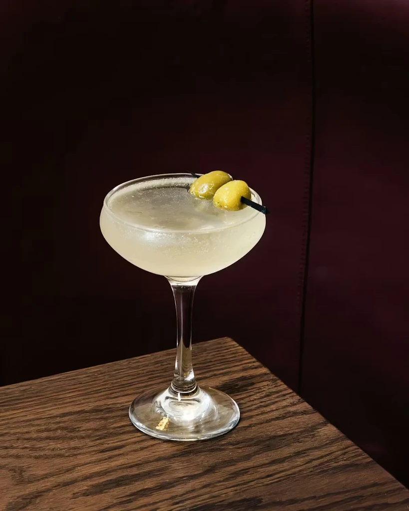 Coupe glass cocktail with two green olives on dark wood.