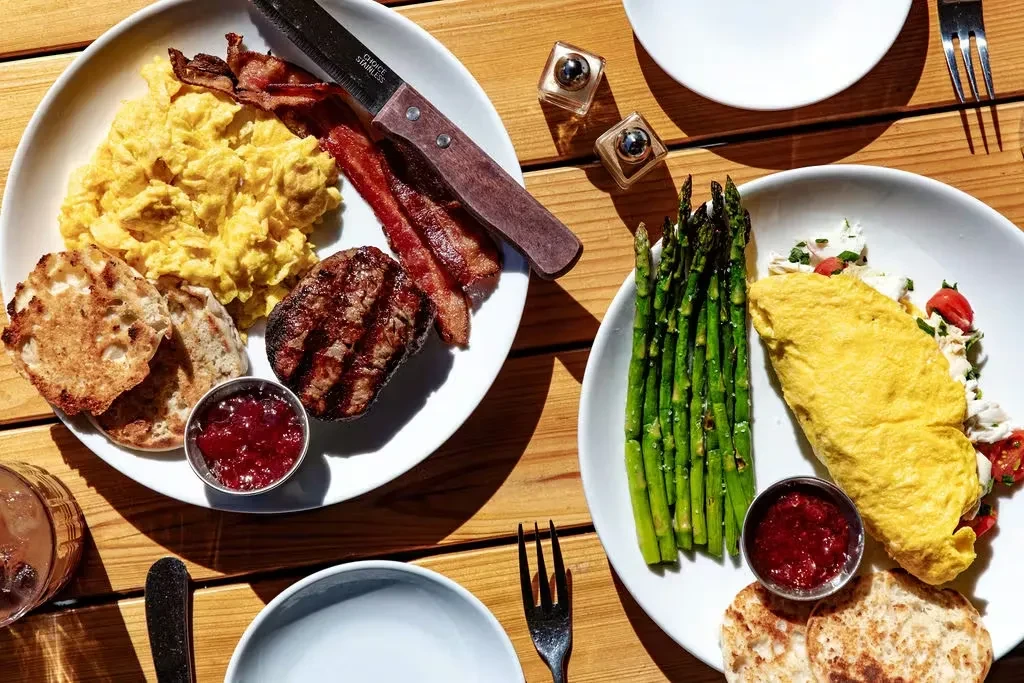 High-angle view: two breakfast plates with eggs, bacon, omelet, muffins, asparagus.