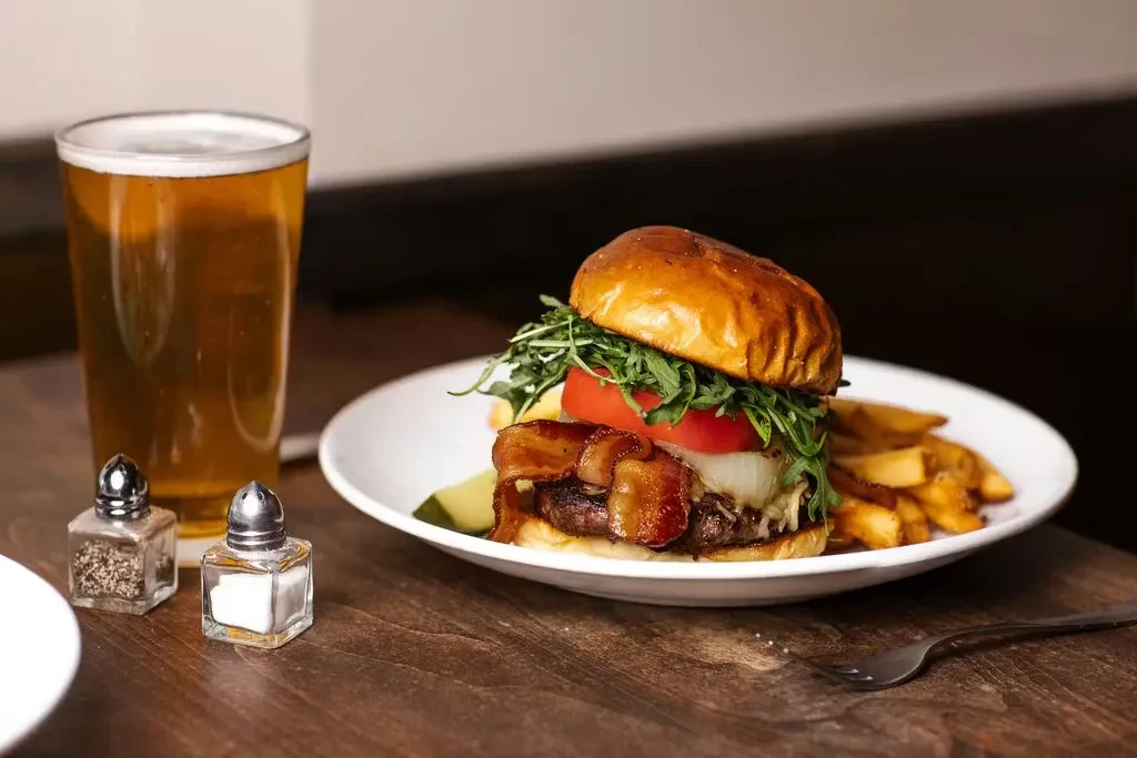 Gourmet burger, fries, pickle, and beer on a dark wooden table.