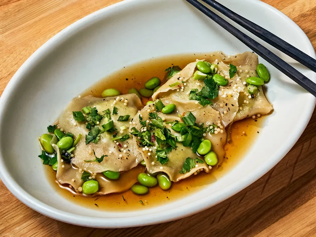 Bowl of dumplings in broth with edamame, green onions, and chopsticks.