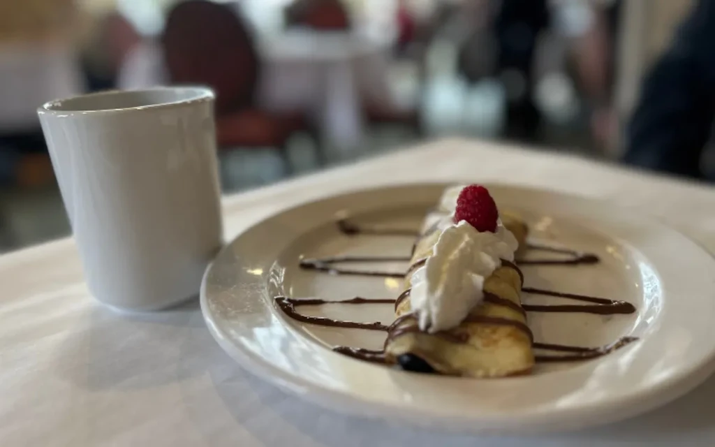 Chocolate-drizzled crepe with whipped cream, raspberry, and a white mug.